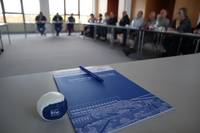 Bfw-Tagungsmappe mit Kugelschreiber im Vordergrund und mehrere Zuh&ouml;rende sitzend im unscharfen Hintergrund